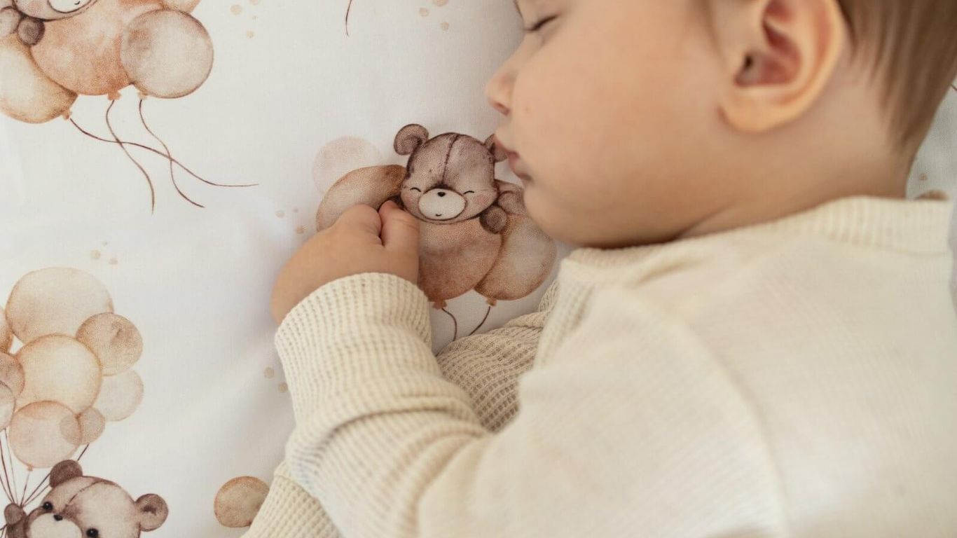 A baby sleeps peacefully on a blanket with teddy bears and balloons.