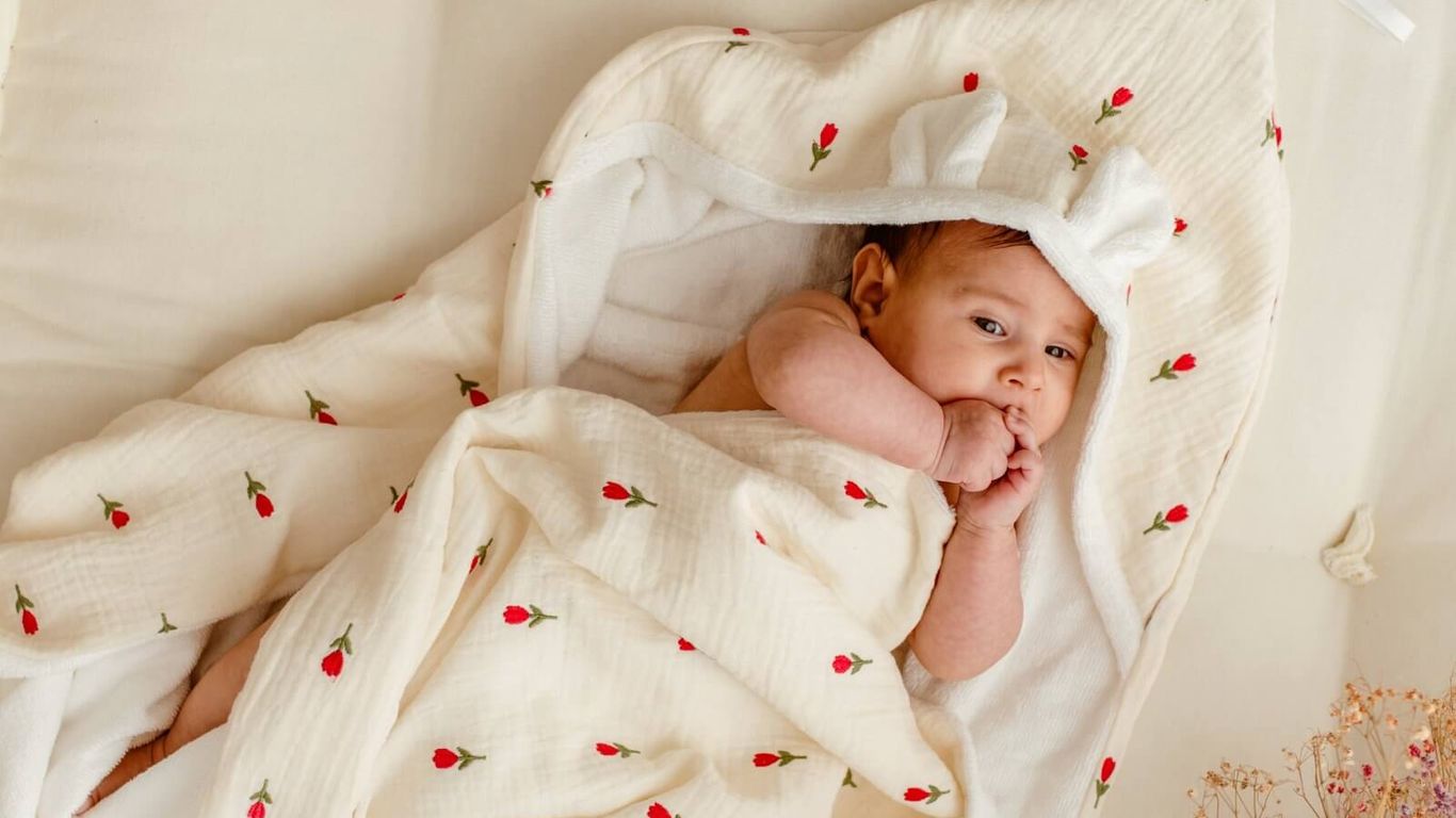 A baby wrapped in a floral blanket with bunny ears on a cream mattress.
