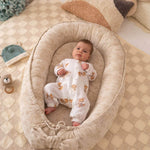Baby in a cozy beige crib with soft pillows and toys in a warm, homey setting.