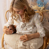 Woman holding a baby and a decorative item in a softly lit room with white curtains and blue flowers.