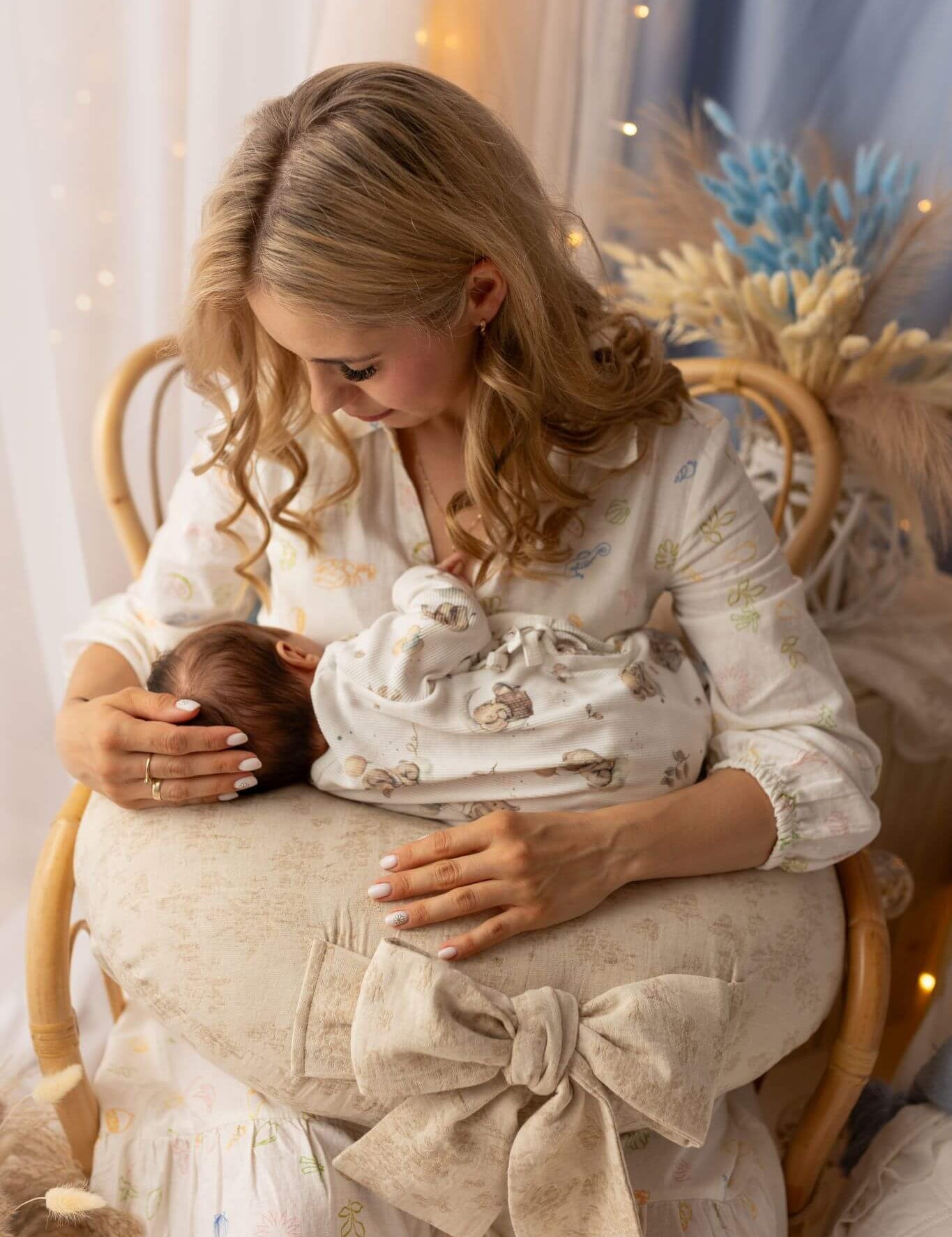 Woman holding a baby and a decorative item in a softly lit room with white curtains and blue flowers.