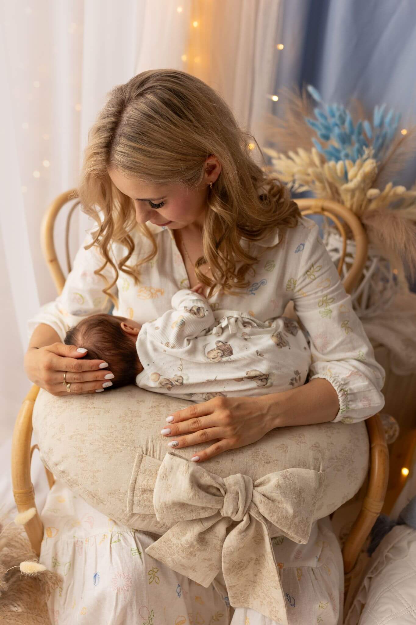 Woman holding a baby and a decorative item in a softly lit room with white curtains and blue flowers.